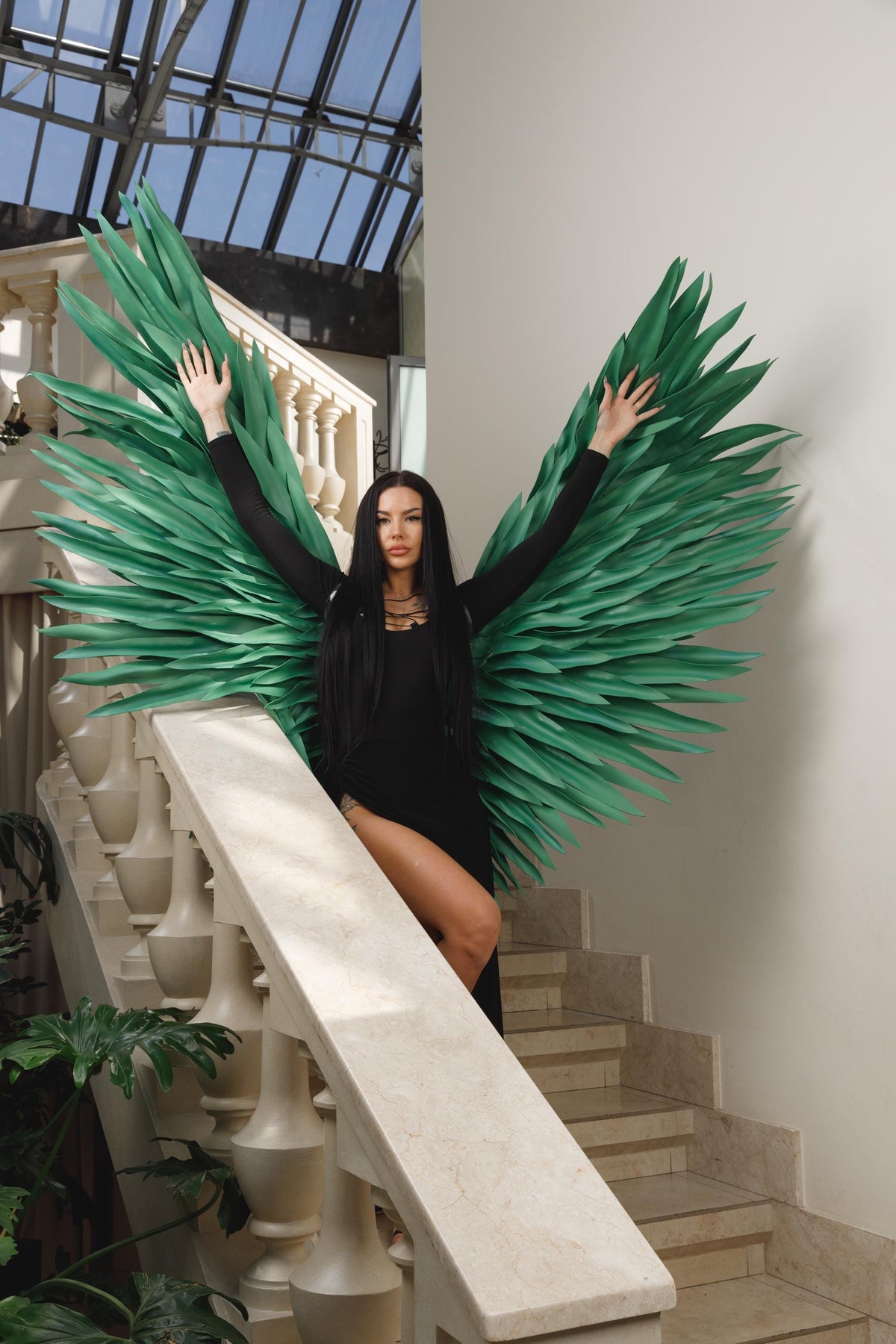 May include: A person wearing a black dress and large green paper wings, posing on a marble staircase. The wings are made of layered, pointed leaves. The background includes a white balustrade, stairs, and a glass ceiling.