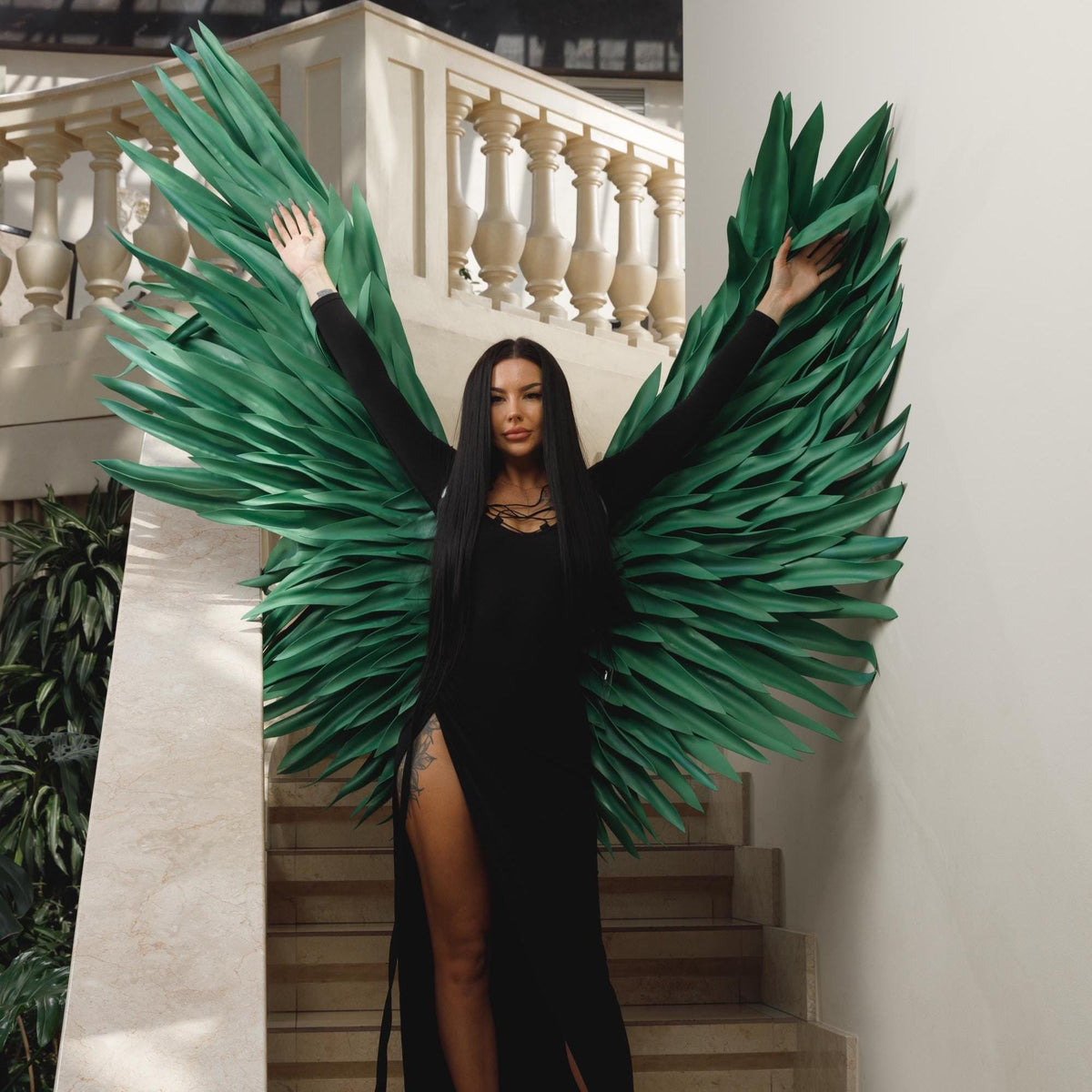 May include: A woman in a black dress with a high slit, posing with large, green, feather-like wings. The wings are attached to a wall, and the woman's arms are outstretched. The background includes stairs and a white balustrade.
