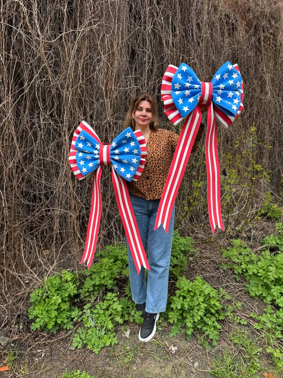 May include: Two large decorative bows with a patriotic theme. The bows feature a blue center with white stars, red and white striped edges, and long red and white striped ribbons. The bows are held up in front of a person.