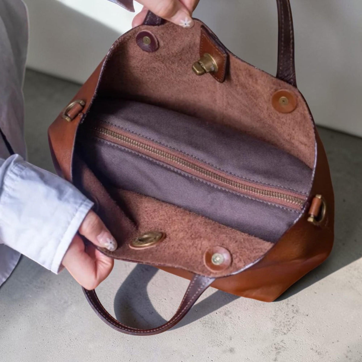 May include: Open brown leather tote bag with a dark brown handle and interior zipper. The bag has a structured shape with metal hardware and a soft, textured interior lining. The bag is held open, showcasing the inside.