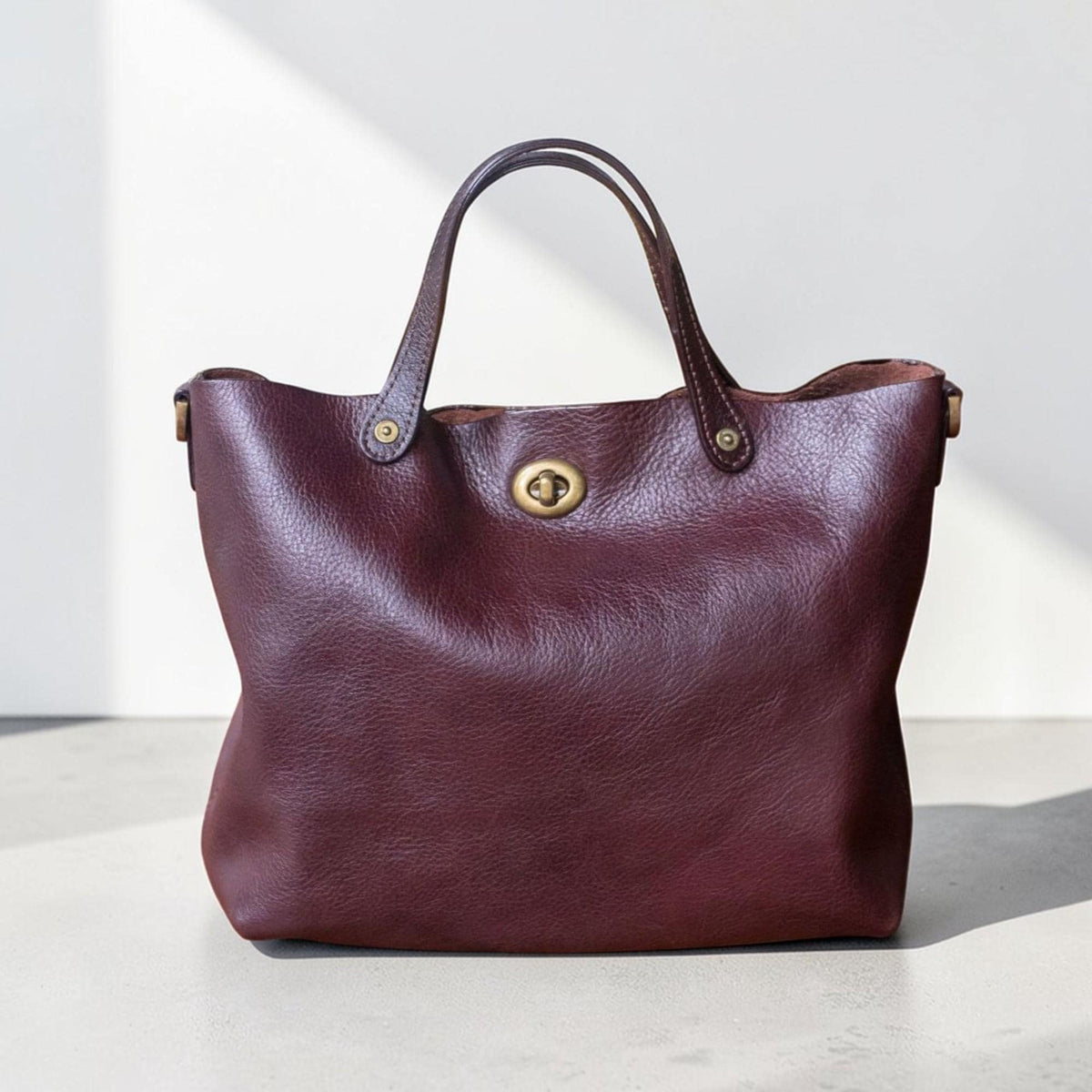 May include: A burgundy leather tote bag with a rounded shape and two handles. The bag has a gold-colored clasp and rivets. The leather has a textured appearance. The bag is designed for carrying items.