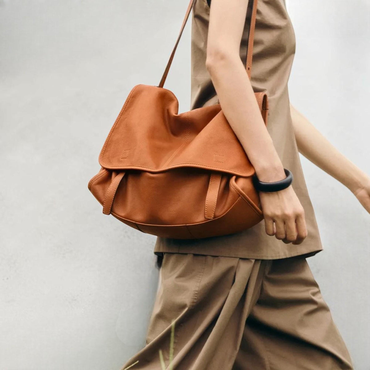 May include: A large, tan leather shoulder bag with a flap closure and adjustable strap. The bag is worn across the body. The person in the image is wearing a tan outfit and a black bracelet.