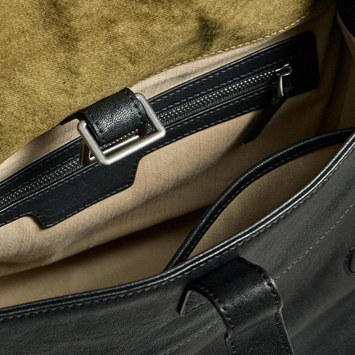 May include: Close-up of a black leather tote bag interior. The bag features a zippered pocket with a silver zipper and a leather strap with a silver buckle. The interior lining is a light tan color, and the bag appears to be well-constructed.