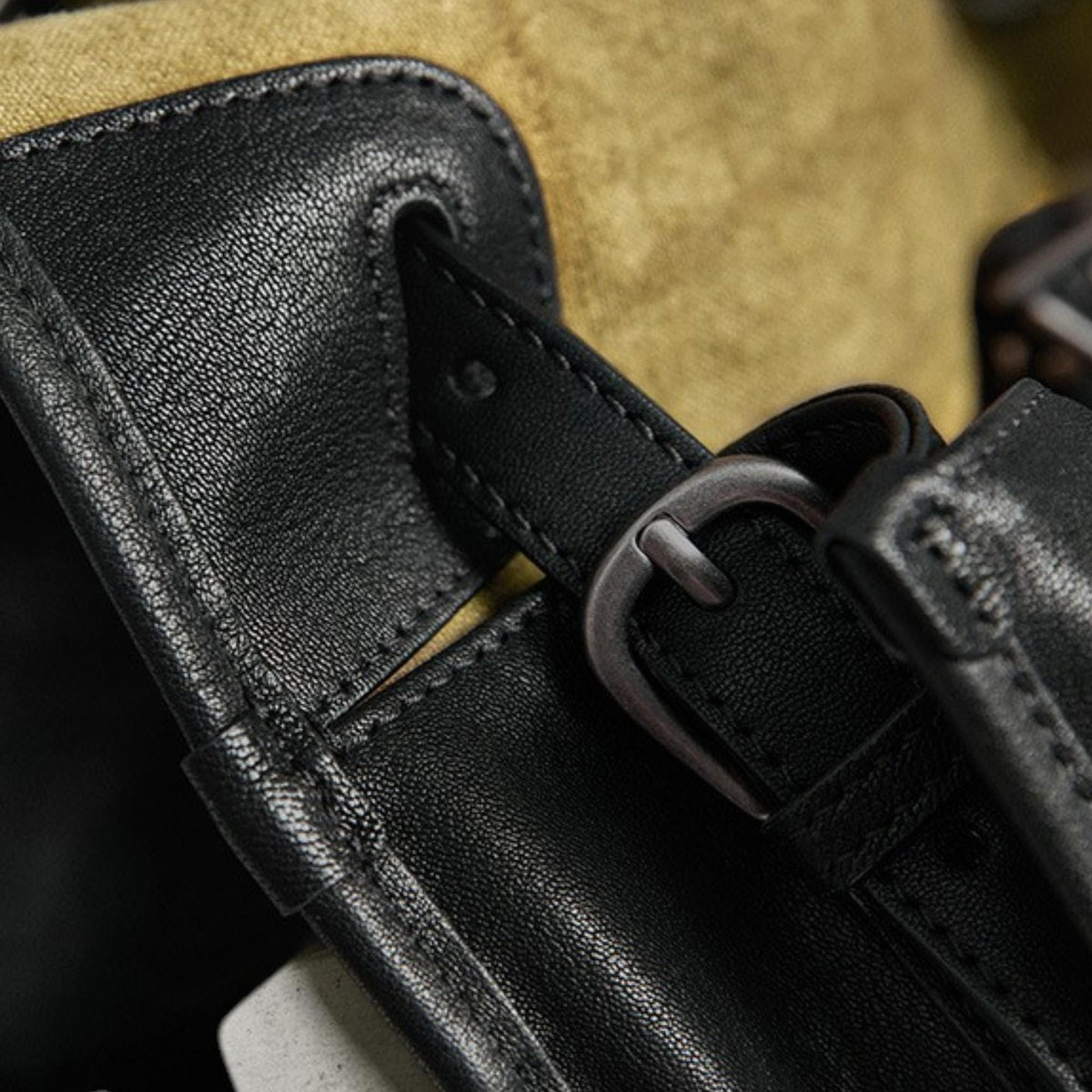 May include: Close-up of a black leather strap with a silver-colored buckle. The strap is stitched to a black leather panel, with a tan fabric visible in the background. The leather has a textured appearance.
