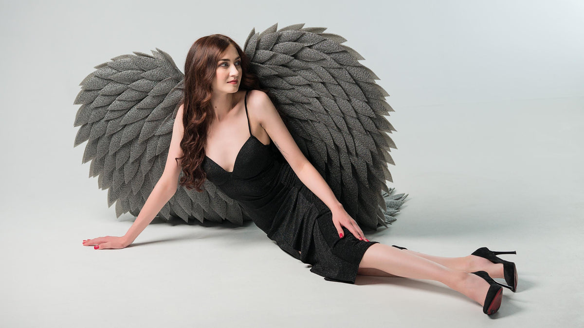 May include: A woman wearing a black dress and high heels poses with large, gray, feathered angel wings. The wings are made of a textured material and are positioned behind her. The background is plain white.