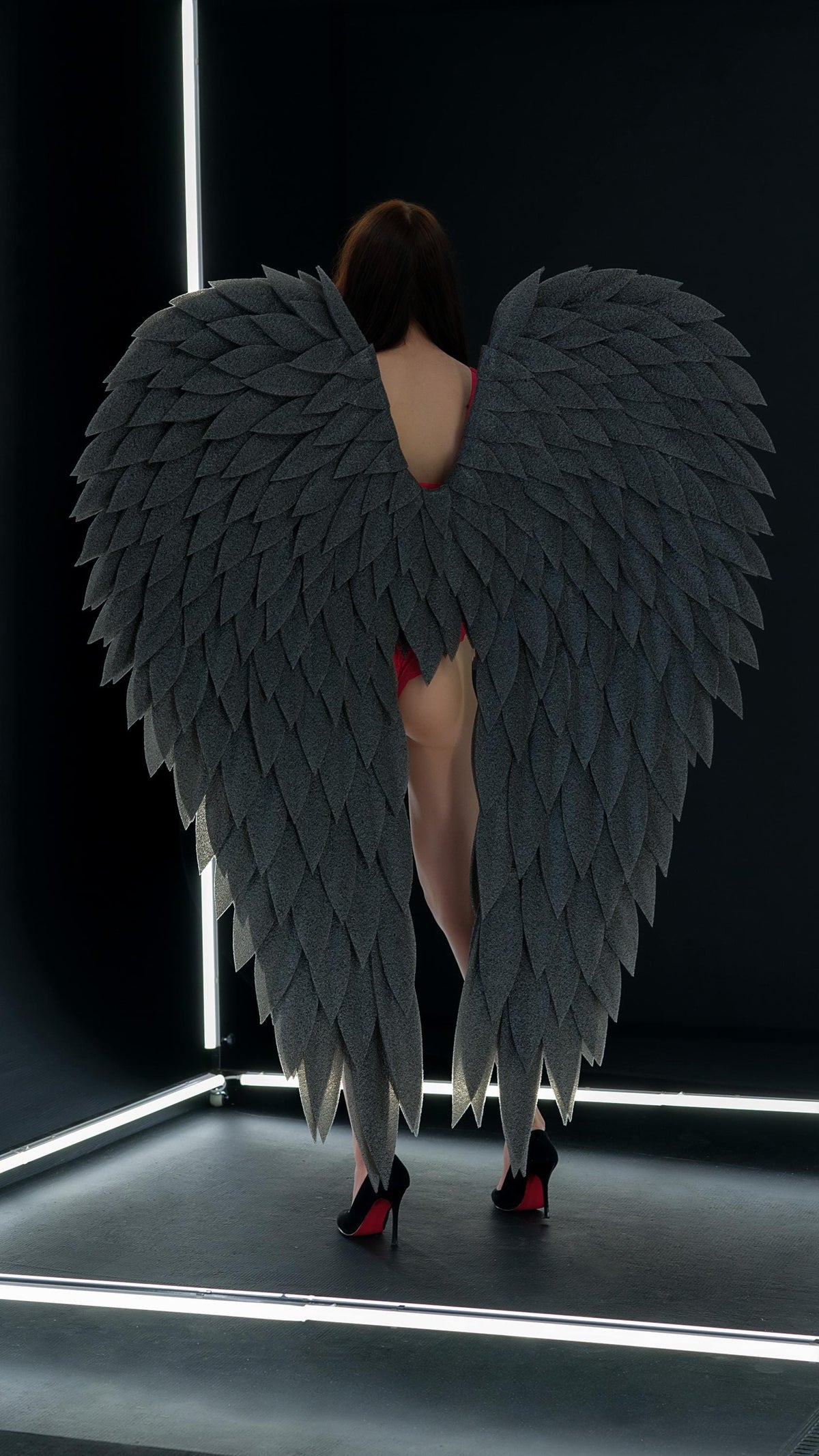 May include: Large, gray, feathered angel wings. The wings are made of many individual, leaf-shaped pieces. The person is wearing black high heels with red soles. The background is black with white neon lights.