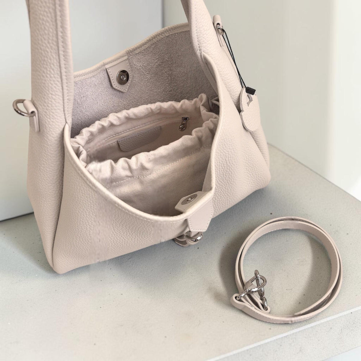 May include: A light beige leather handbag with a drawstring inner pouch. The bag has a shoulder strap and a detachable strap. The bag is open, revealing the interior. The bag is sitting on a light-colored surface.