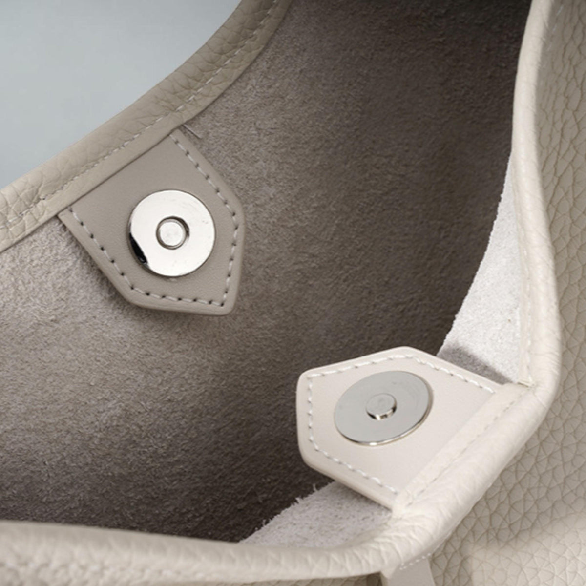 May include: Close-up of a light gray leather handbag interior. The bag features a magnetic closure with a silver metal disc. The interior is a soft, textured gray, and the edges are stitched with a contrasting thread.