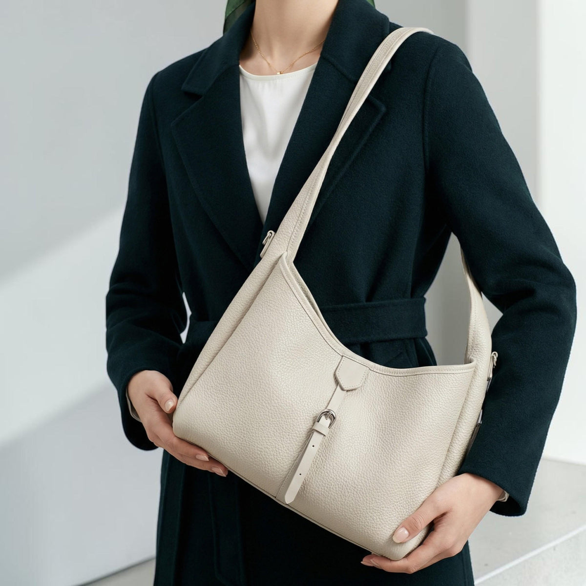 May include: A cream-colored leather shoulder bag with a curved design. The bag features a silver buckle closure and a long strap. The bag is held by a person wearing a dark green coat.