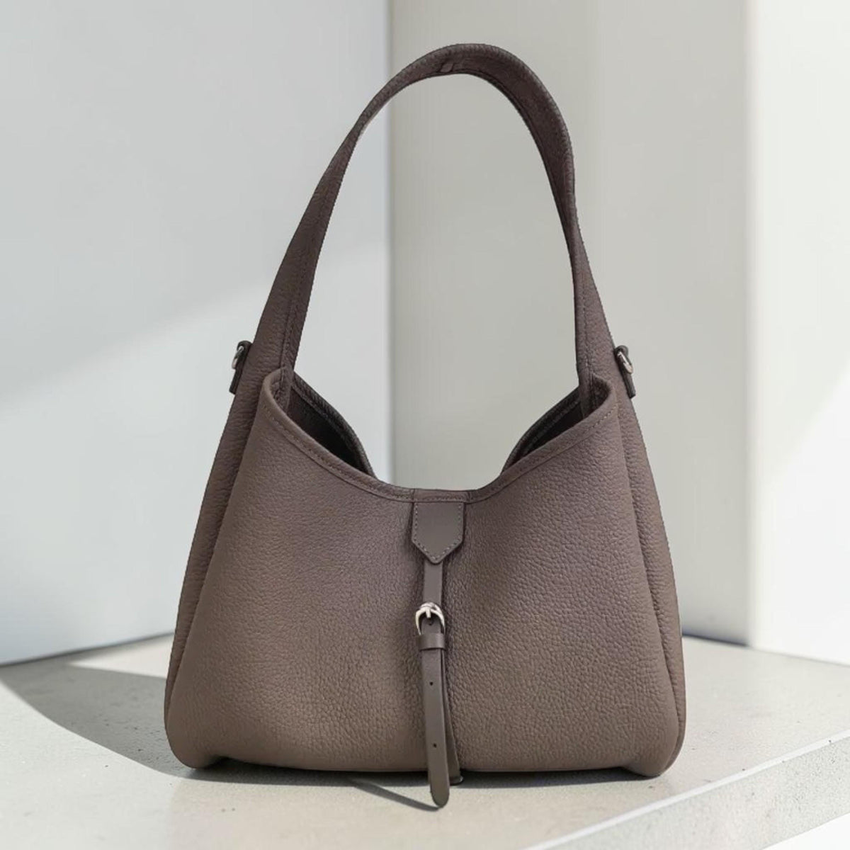May include: A taupe-colored handbag with a single shoulder strap. The bag has a rounded shape with a small strap and buckle detail at the front. The leather has a textured appearance.