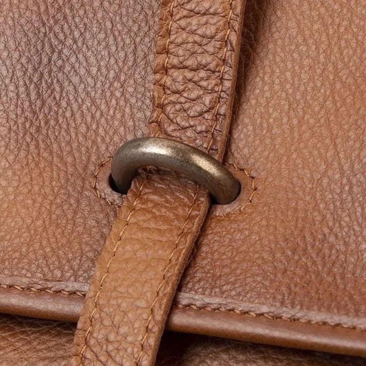 May include: Close-up of a brown leather bag strap with a metal ring. The strap is stitched and has a textured surface. The metal ring is a dark, muted silver color.