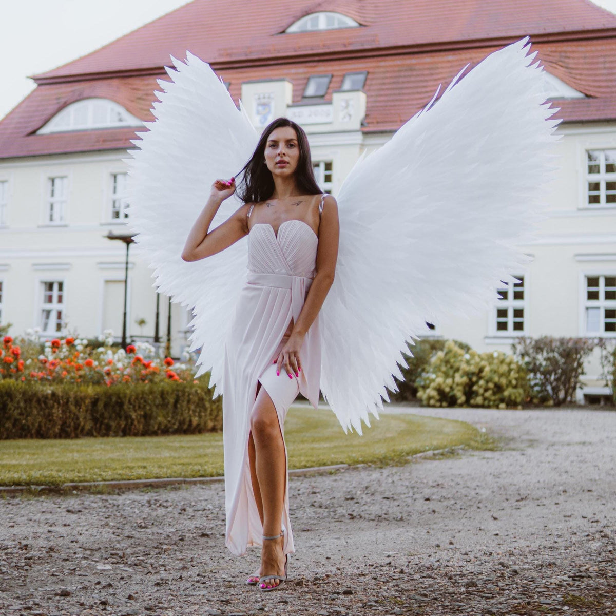 May include: A woman wearing a light pink dress and large white feathered angel wings stands in front of a building with a red roof. The dress has a high slit and the woman is wearing high heels.