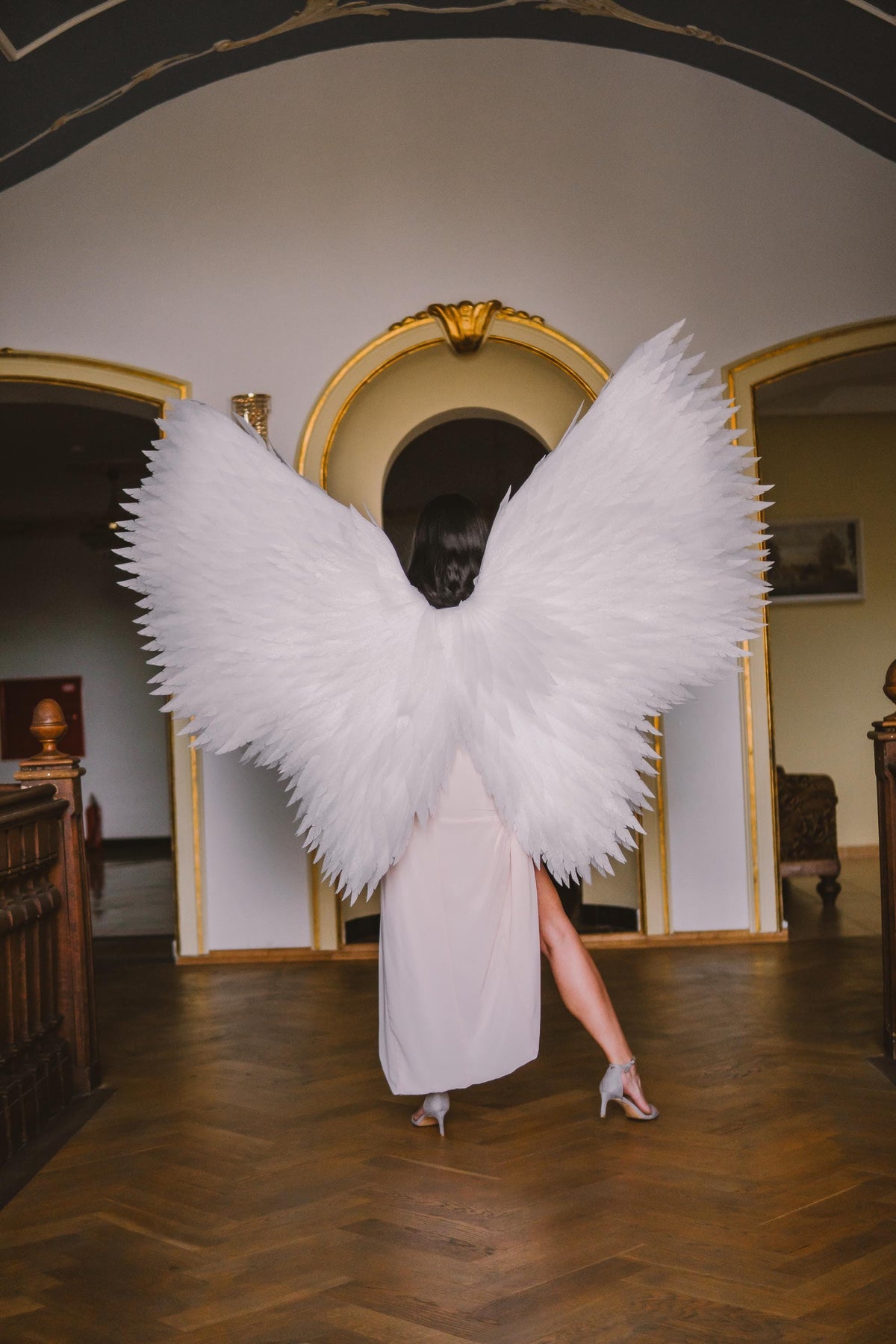 May include: A person wearing large, white feathered angel wings. The wings are the focal point, with a detailed texture. The person is wearing a light-colored dress and heels, standing in a room with ornate architectural details and a wooden floor.