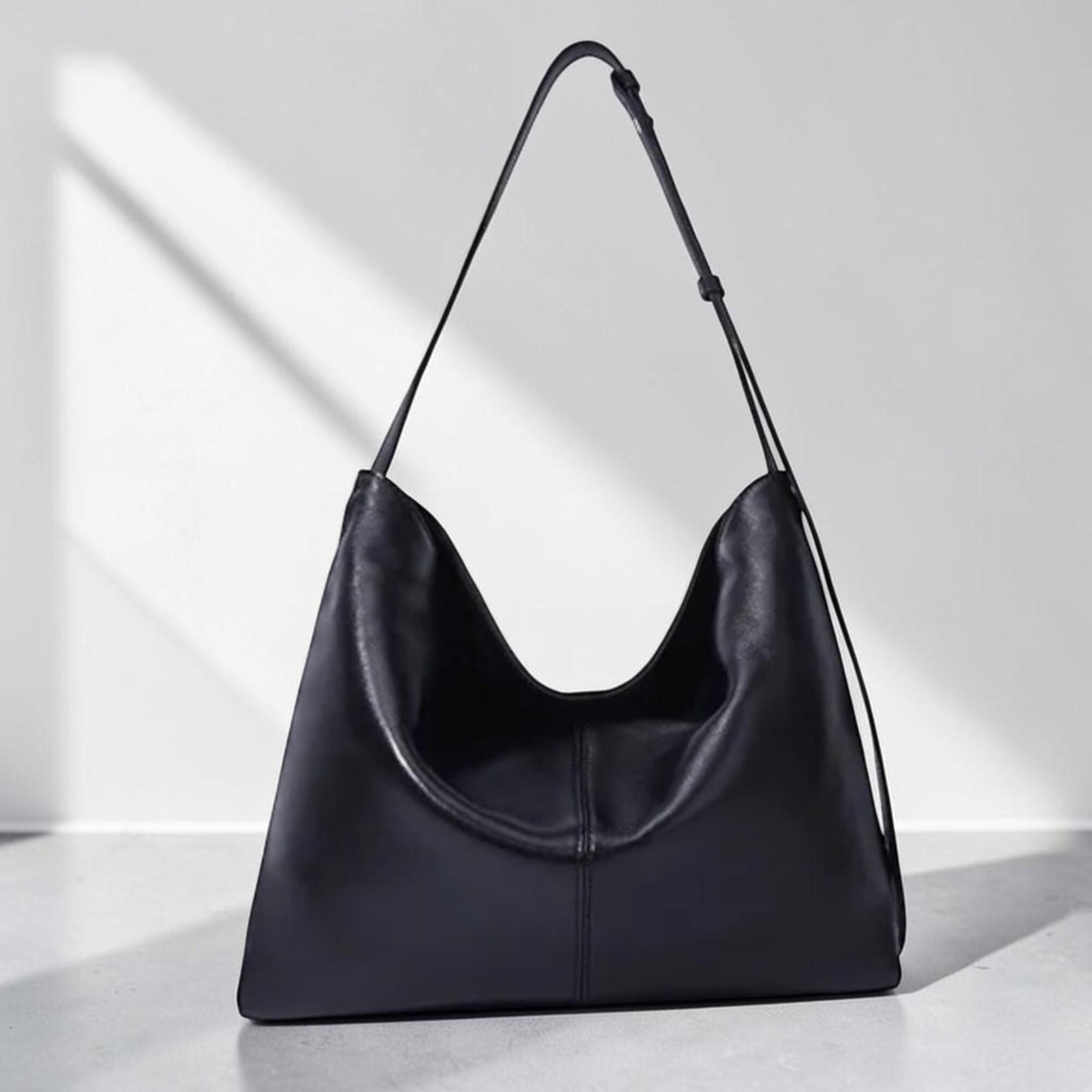 May include: A black leather shoulder bag with a long strap. The bag has a curved, open top and a triangular shape. The bag is made of smooth leather and has a minimalist design. The bag is sitting on a white surface.