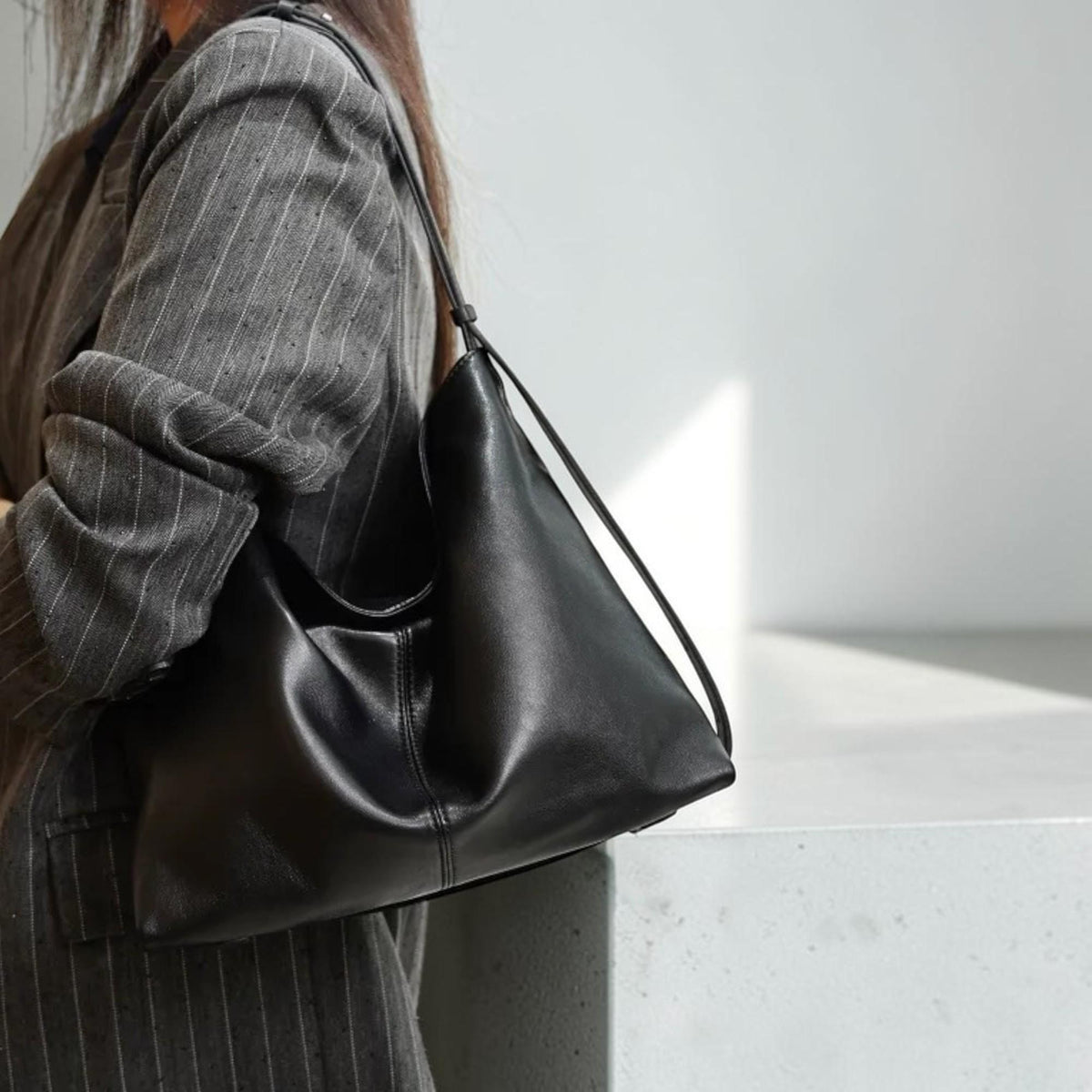 May include: A black leather shoulder bag with a minimalist design. The bag has a single strap and a soft, unstructured shape. The bag is worn over the shoulder and appears to be empty. The background is a neutral white and gray.