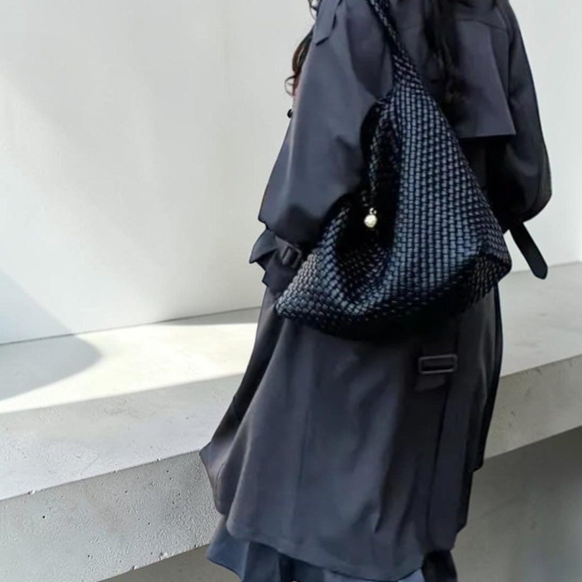 May include: A dark navy blue woven shoulder bag with a pearl charm. The bag is carried over the shoulder and hangs against a dark navy blue trench coat. The bag has a woven pattern.