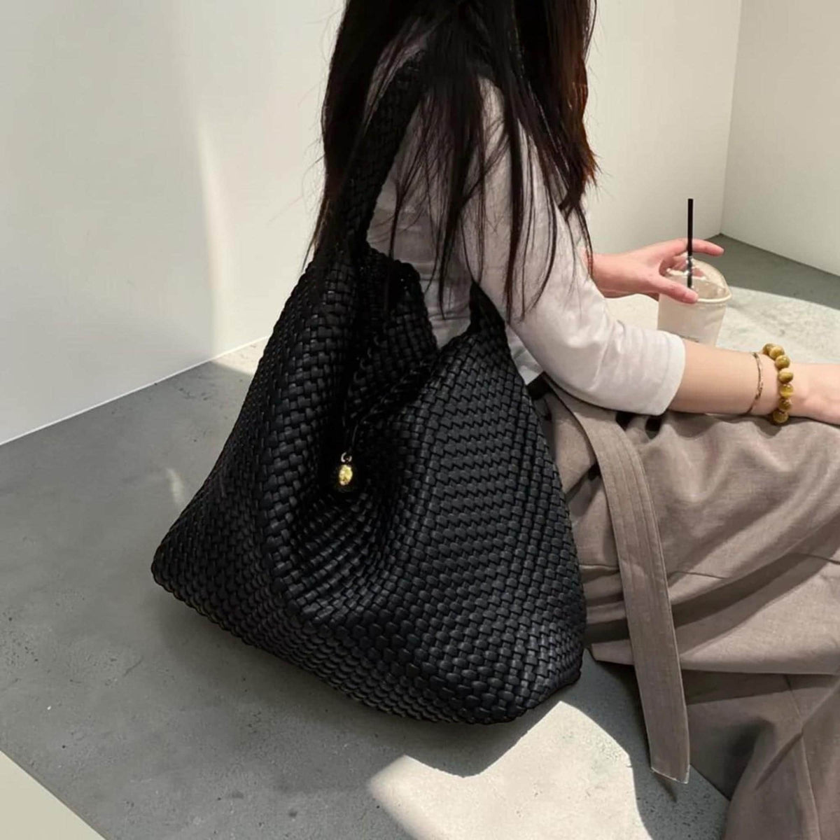 May include: A black woven tote bag with a shoulder strap. The bag has a textured, basket-weave design and a small gold-colored charm. The bag is large and appears to be empty. The bag is a neutral accessory.