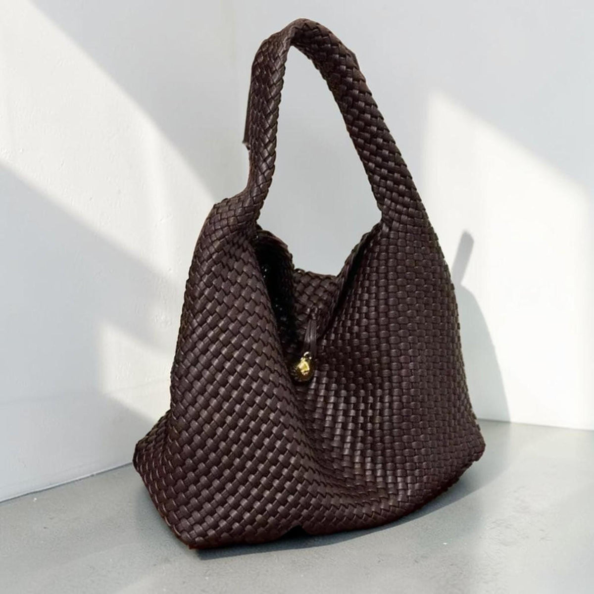 May include: A dark brown woven handbag with a single braided handle. The bag has a slouchy, hobo-style shape and a gold-colored clasp. The weave pattern is tight and detailed, creating a textured surface.