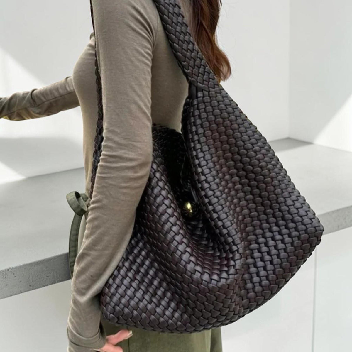 May include: A dark brown woven leather shoulder bag with a large, slouchy design. The bag features a wide strap and a gold-colored clasp. The woven pattern creates a textured appearance. The bag is worn over the shoulder.