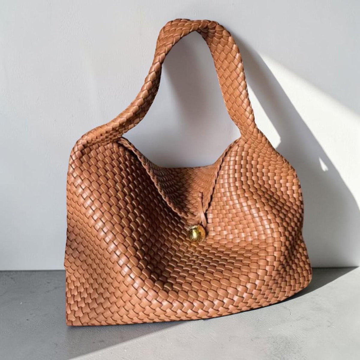 May include: A woven brown handbag with a single handle. The bag features a large, rounded shape with a woven texture and a gold-colored button closure. The bag is made of a soft, flexible material.