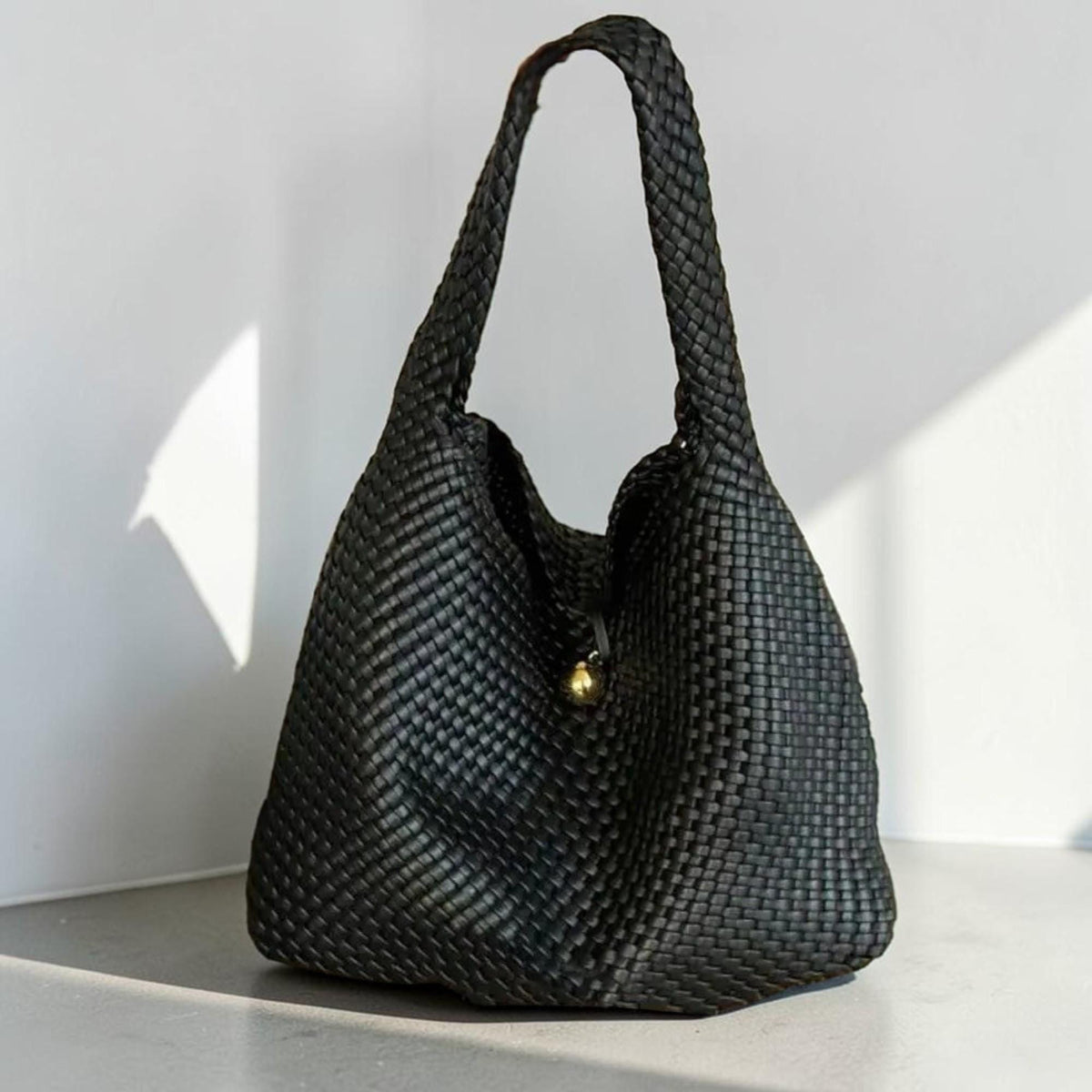 May include: A black woven tote bag with a single shoulder strap. The bag has a rounded shape and a gold-colored clasp. The weave pattern is visible throughout the bag, creating a textured appearance. The bag is sitting on a light-colored surface.