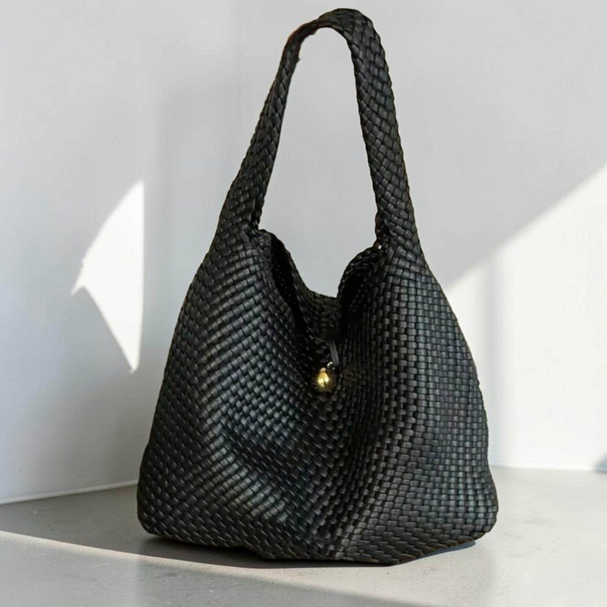 May include: A black woven tote bag with a single shoulder strap. The bag has a rounded shape and a gold-colored clasp. The weave pattern is visible throughout the bag, creating a textured appearance. The bag is sitting on a light-colored surface.