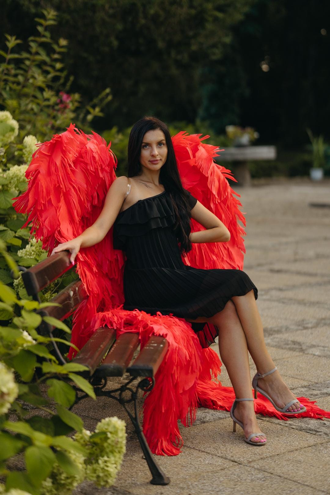 May include: A woman wearing a black dress and red feathered angel wings sits on a park bench. The wings are large and vibrant, creating a striking contrast against the dark dress. The scene is set outdoors with greenery in the background.