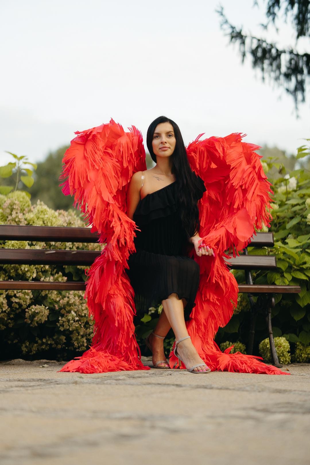 May include: A woman wearing a black dress and red feathered wings sits on a wooden bench. The wings are large and vibrant, creating a striking contrast against the dark dress. The setting is outdoors, with greenery in the background.