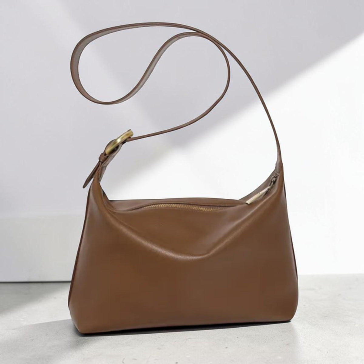 May include: A smooth brown shoulder bag with a curved silhouette, featuring a top zipper closure and a long, adjustable strap attached with a circular brass-toned metal buckle. The bag has a clean, structured design with a soft, matte finish.