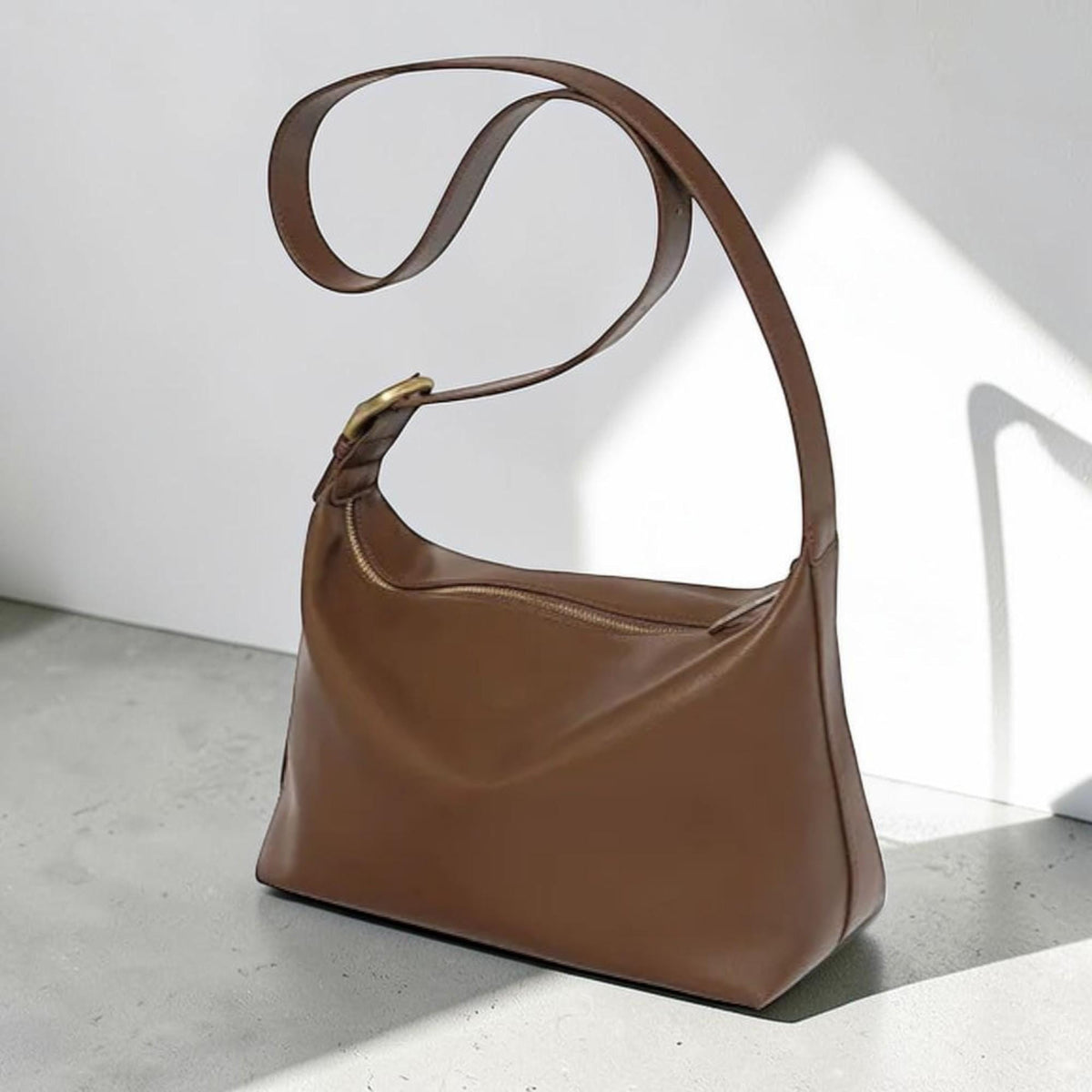May include: A smooth brown shoulder bag with a curved silhouette and a single wide strap. The bag features a top zipper closure and a metallic gold-toned buckle detail on the strap, providing a clean and structured appearance for daily use.