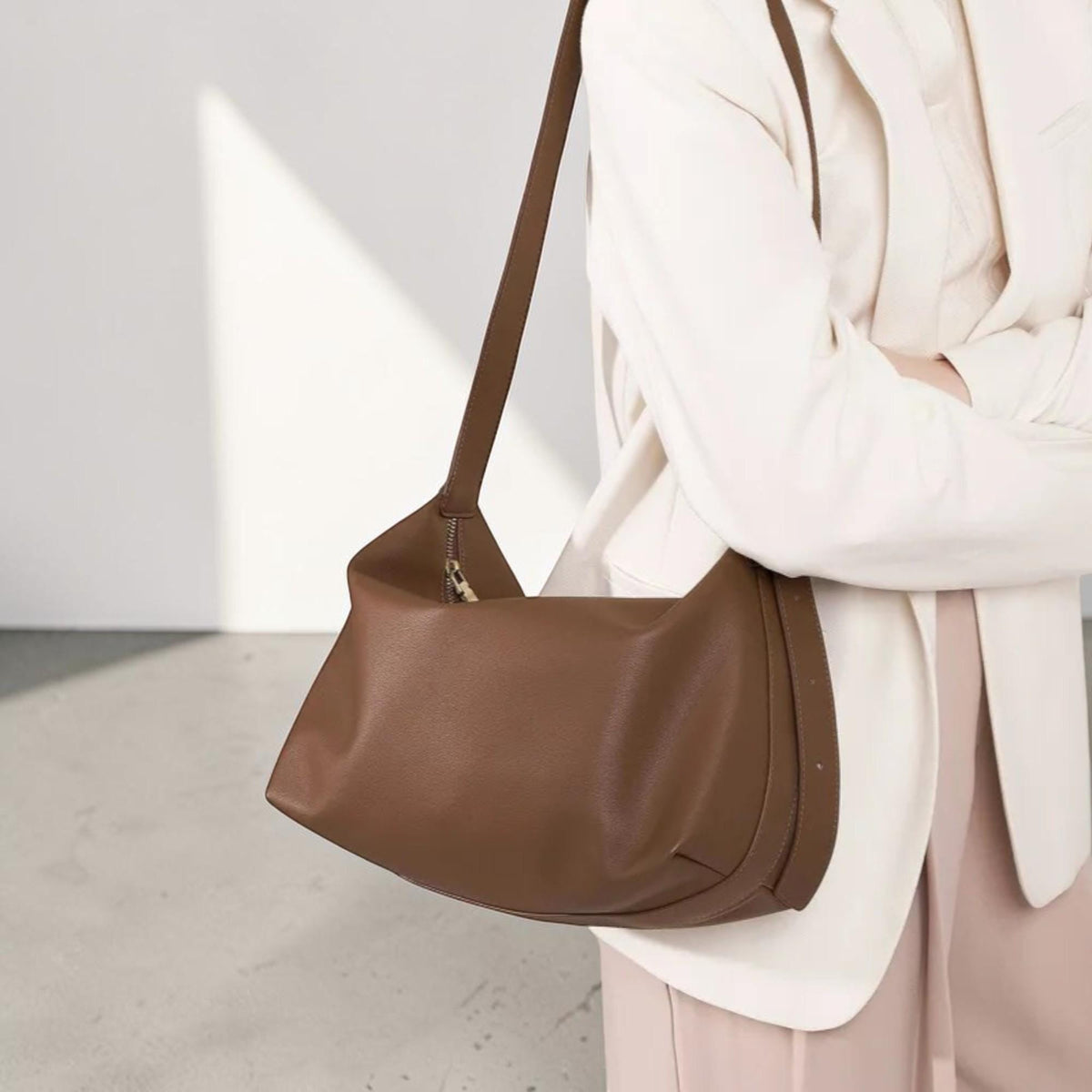 May include: A smooth brown shoulder bag with a curved silhouette, featuring a single long strap and a top zipper closure. The bag has a structured base and a soft, matte finish, providing a clean and functional design for carrying daily essentials.