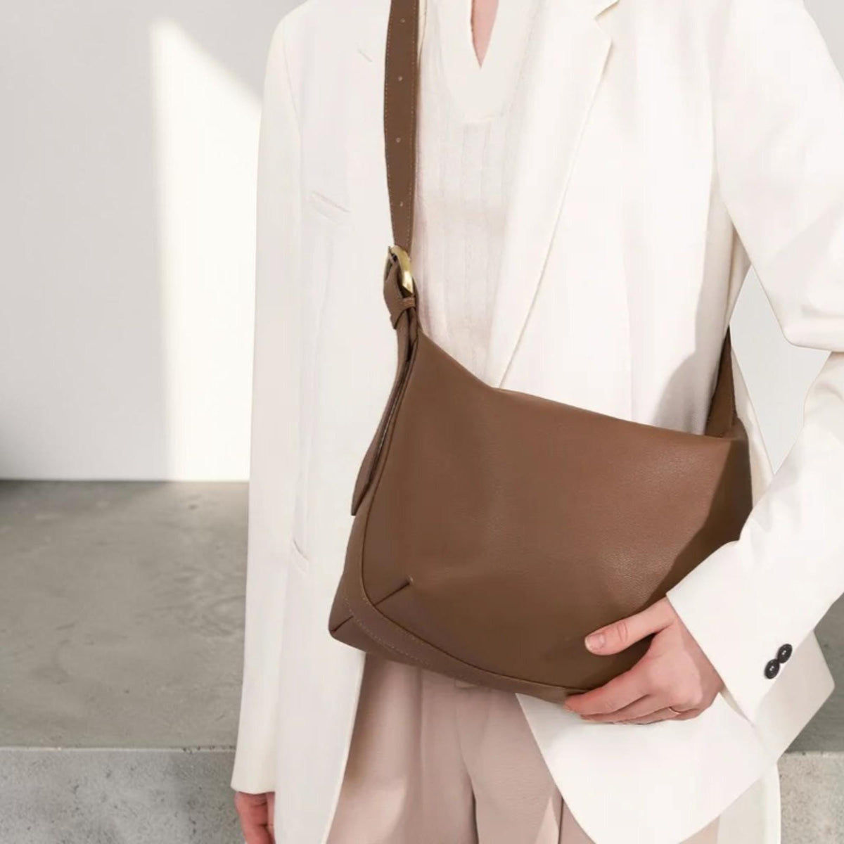 May include: A brown shoulder bag with a smooth texture and a long adjustable strap featuring a gold-toned metal buckle. The bag has a curved bottom edge and a simple, clean design, worn over a white blazer and a matching white top.