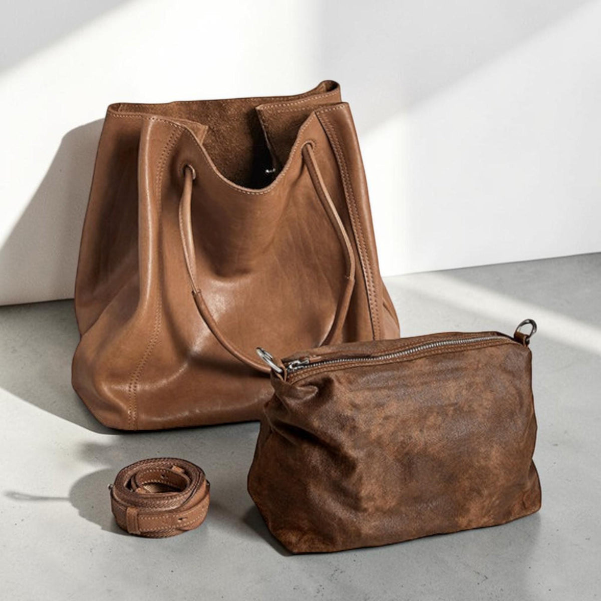 May include: A set of three brown leather bags. The largest is a tote bag with a drawstring closure. A smaller, rectangular bag with a zipper closure and a rolled-up strap are also included. The bags are all a similar shade of brown.