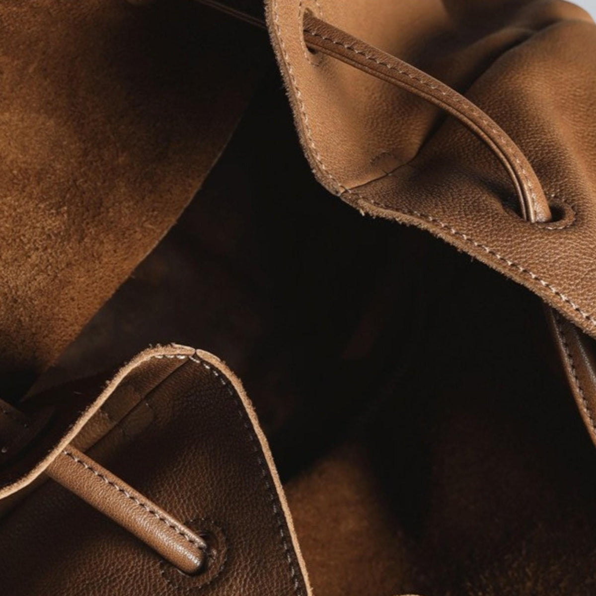 May include: Close-up of a brown leather bag with a drawstring closure. The bag's interior is visible, revealing a soft, suede-like texture. The leather appears to be distressed, with visible stitching and a natural, earthy tone.