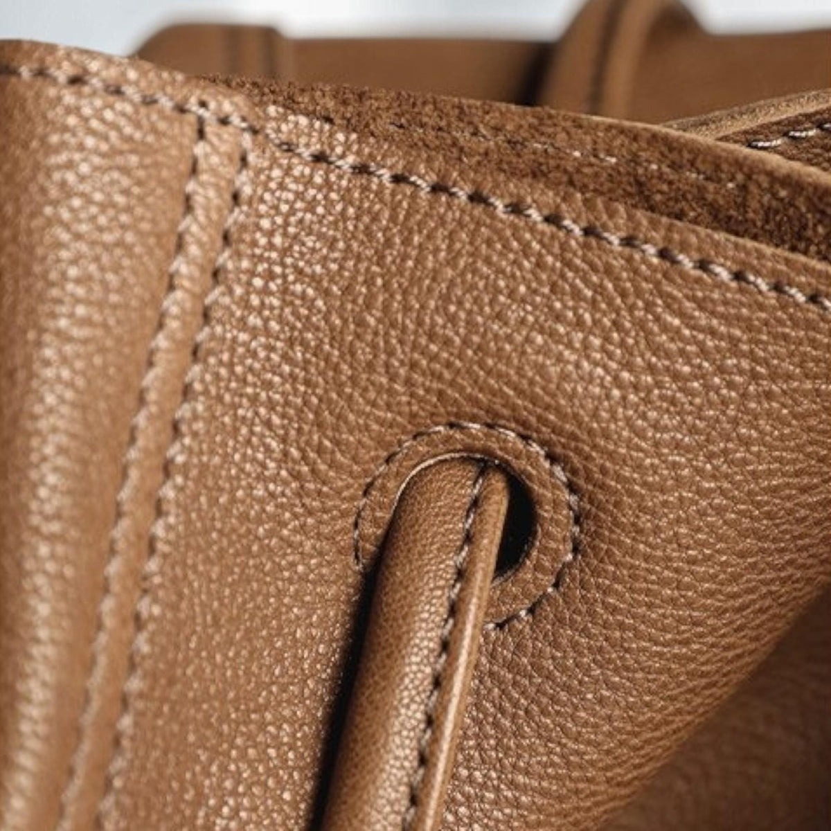 May include: Close-up of a brown leather handbag. The image highlights the texture of the leather, the stitching, and a handle detail. The leather appears to be a rich, warm brown color, with visible grain and subtle variations in tone.