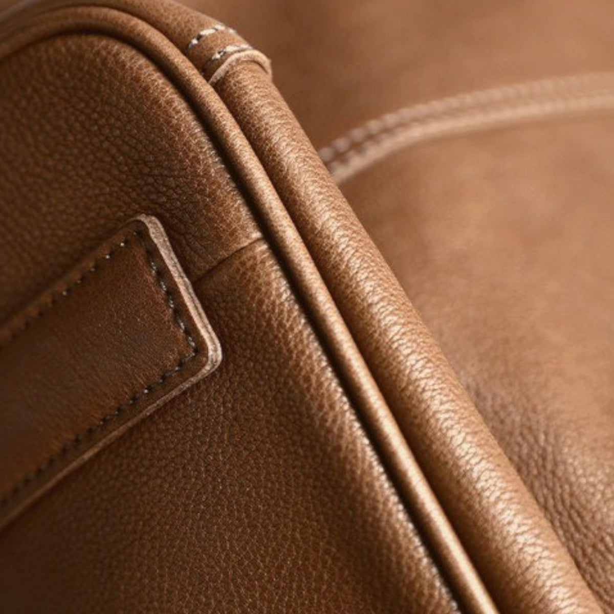May include: Close-up of brown leather with visible grain and stitching. The leather appears to be part of a bag or other accessory, showcasing the texture and craftsmanship. The stitching is a lighter color, providing contrast.