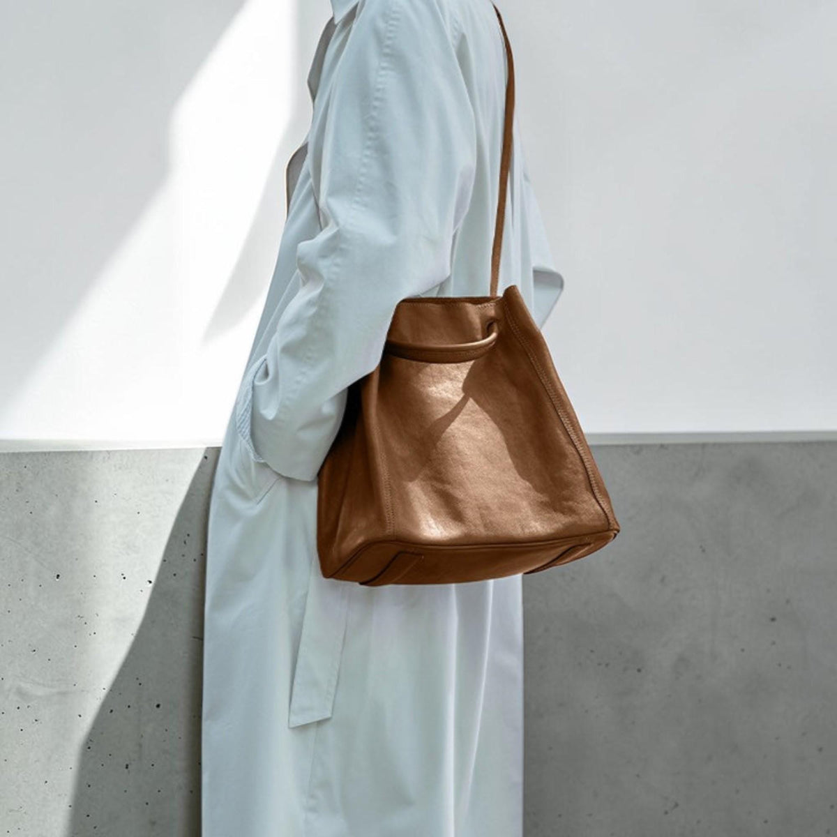 May include: A brown leather tote bag with a shoulder strap. The bag has a rectangular shape with a rounded top handle. The bag is carried by a person wearing a white coat.