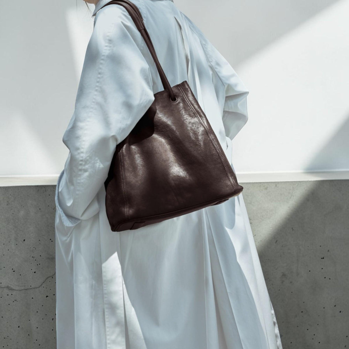 May include: A dark brown leather tote bag with a long shoulder strap. The bag has a rectangular shape with a slightly tapered design. The leather appears to have a textured finish. The bag is carried over the shoulder.