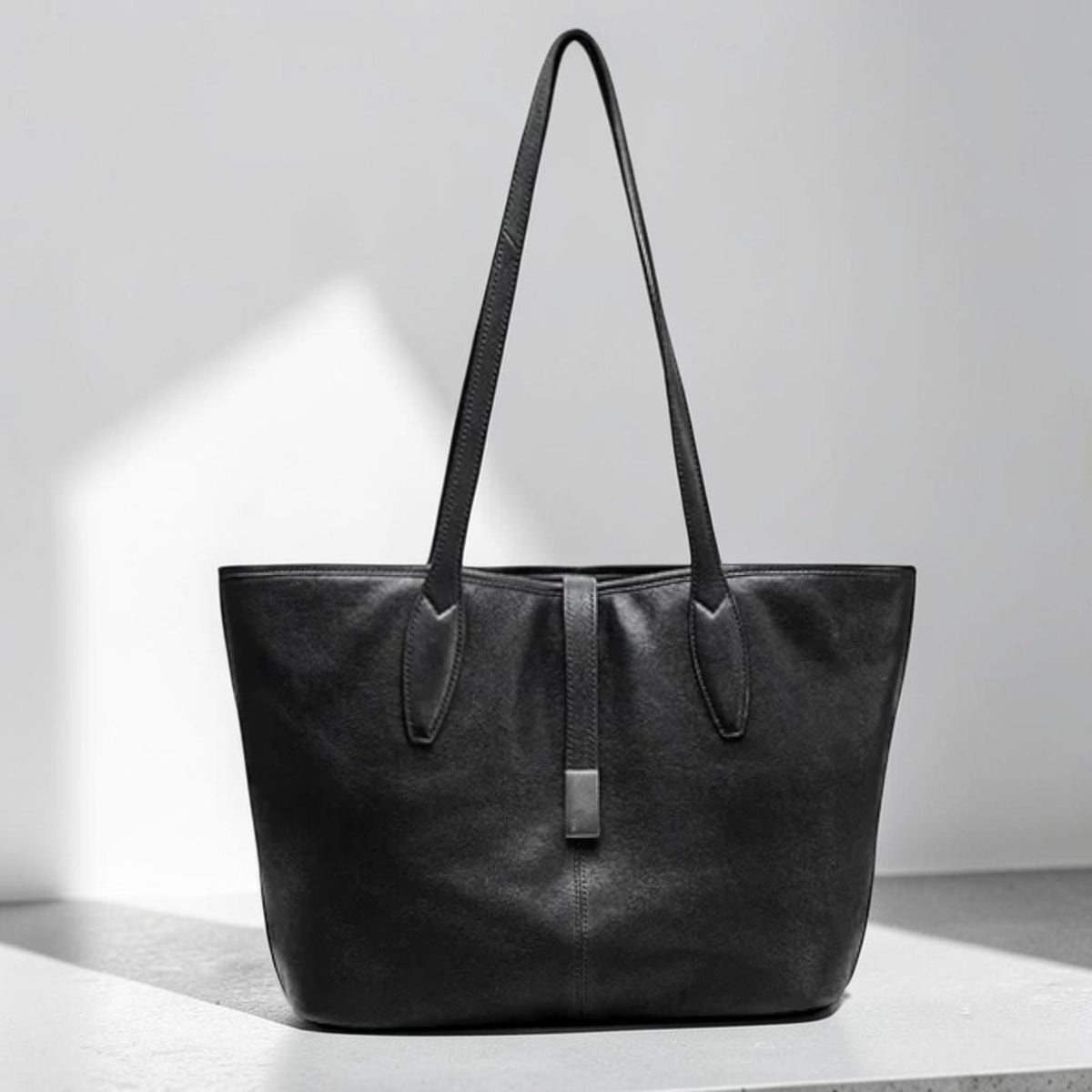 May include: A black leather tote bag with long handles. The bag has a rectangular shape with a vertical strap and a metal clasp in the center. The bag is made of a smooth, matte leather material.