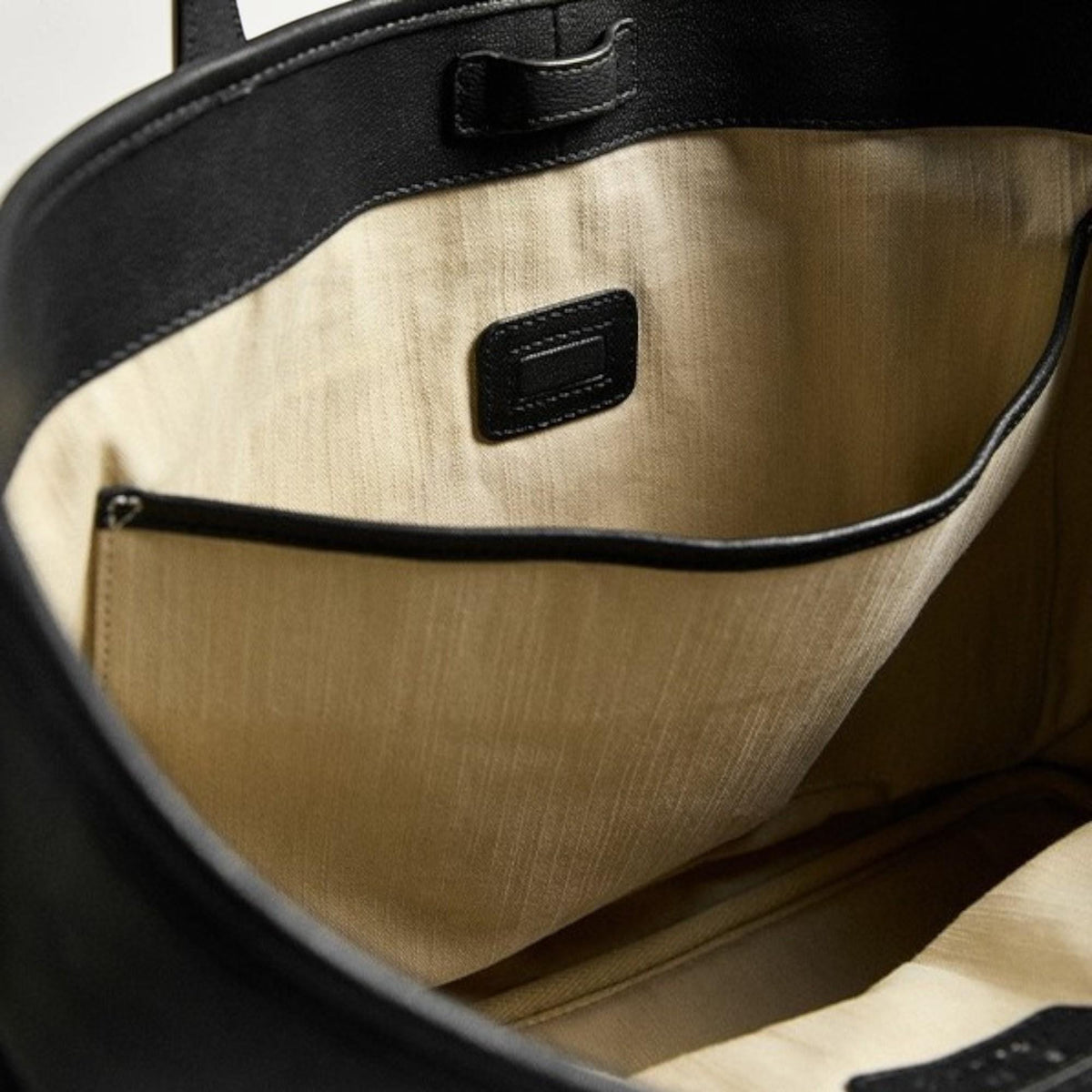 May include: Black leather tote bag with a beige fabric interior. The bag has a small pocket and a leather patch inside. The top of the bag has a handle and a small leather strap.