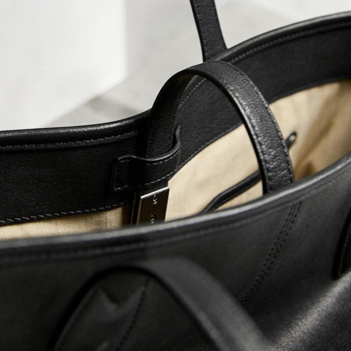 May include: Close-up of a black leather tote bag. The bag features a structured design with a top handle and a silver-colored metal clasp. The interior lining is a light beige color, contrasting with the black exterior.