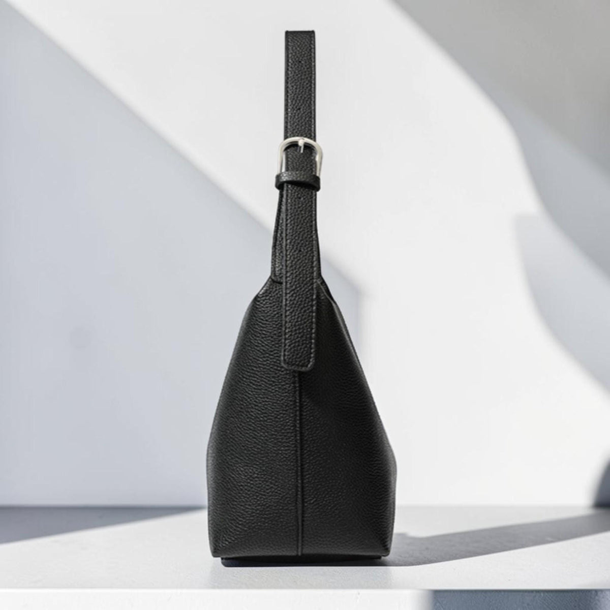 May include: A black leather handbag with a single handle and a silver buckle. The bag has a structured, trapezoidal shape and a textured surface. The handle is attached to the top of the bag with a silver buckle.