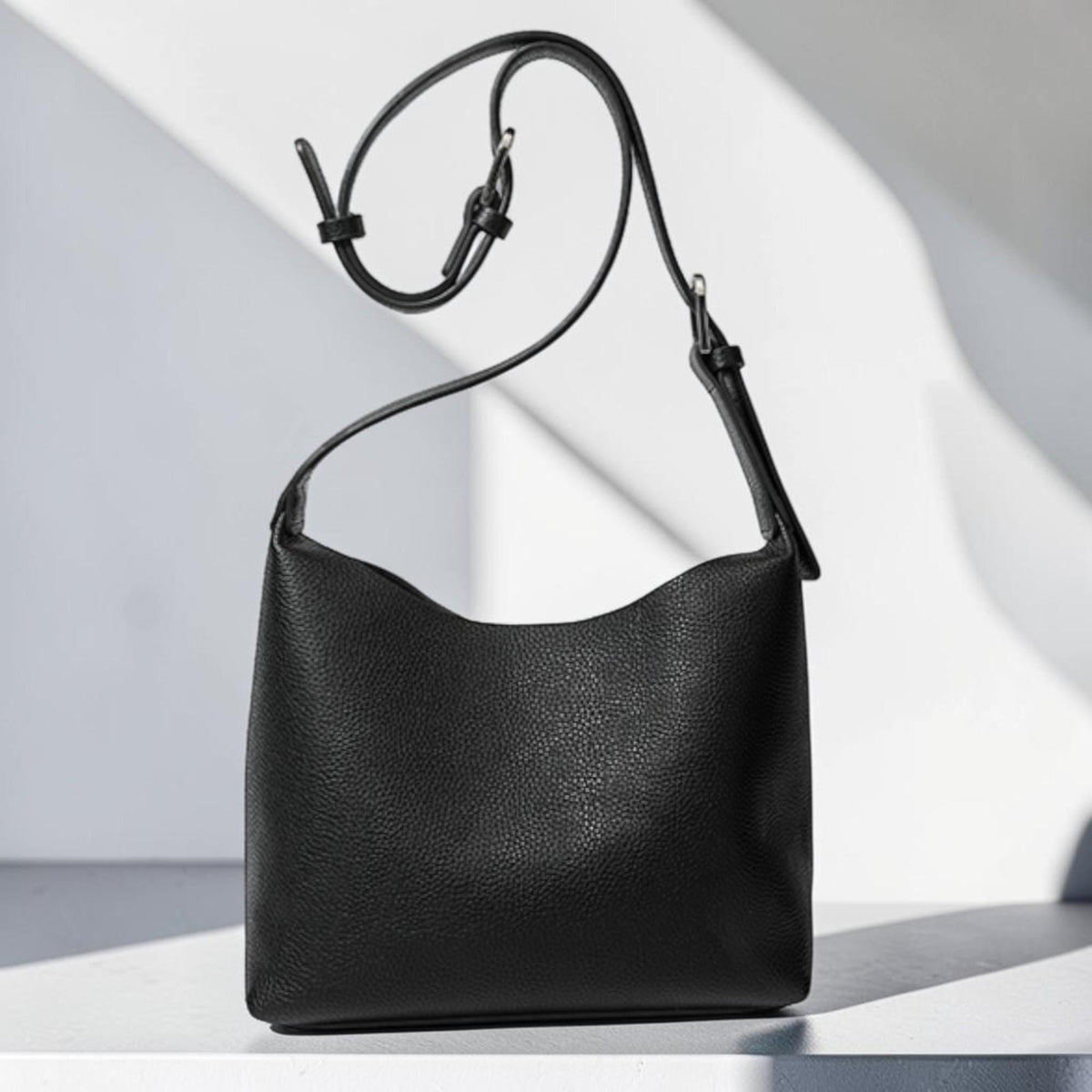 May include: A black leather shoulder bag with a curved top edge and adjustable strap. The bag has a textured surface and a simple, minimalist design. The strap is thin and attaches to the bag with metal hardware.