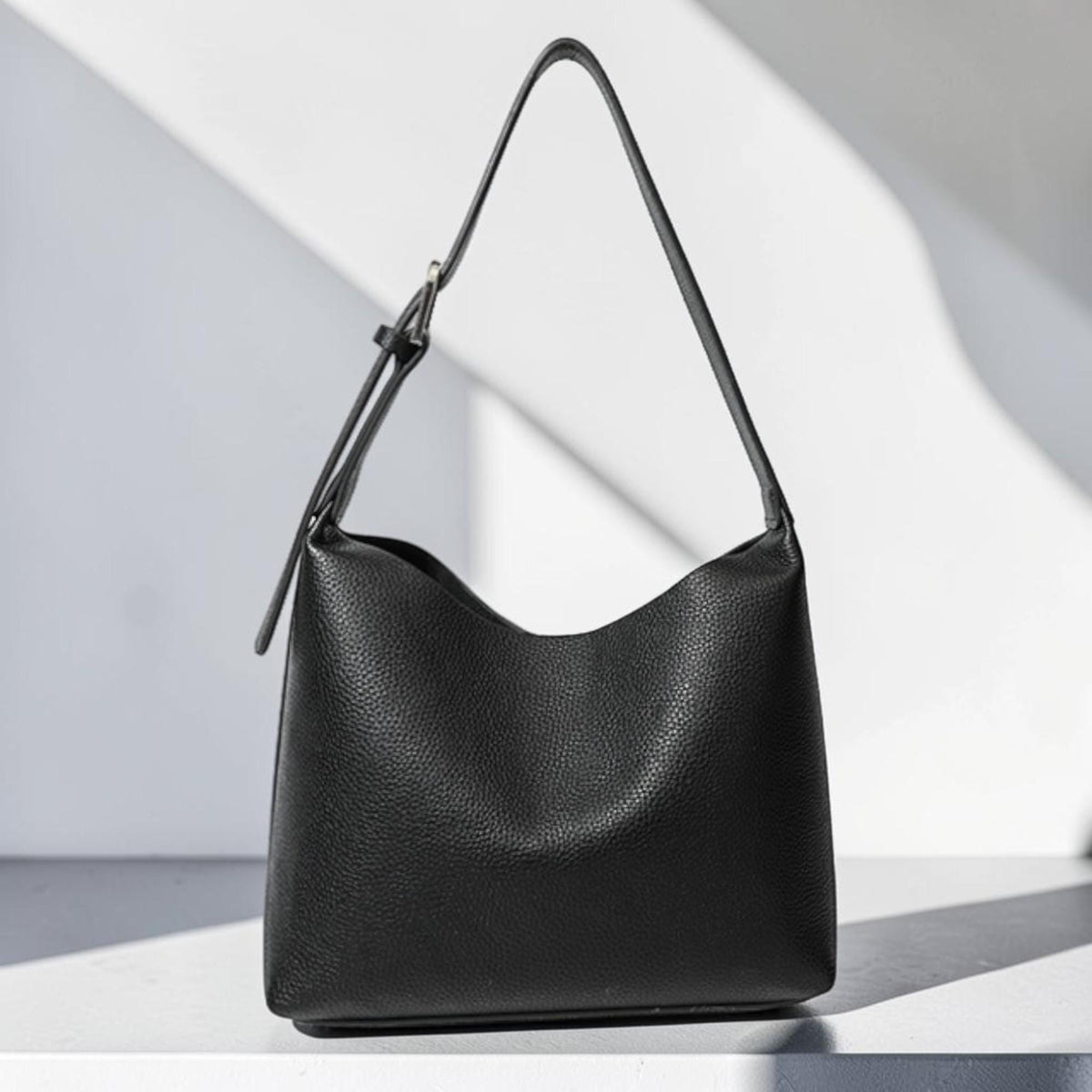 May include: A black leather shoulder bag with a single strap. The bag has a curved top and a textured surface. The strap is adjustable with a silver-colored buckle. The bag is sitting on a white surface.