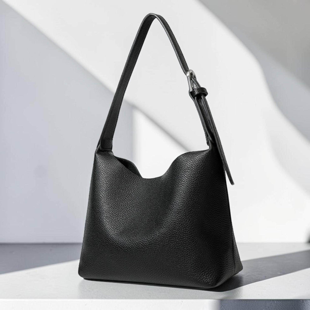 May include: A black shoulder bag with a single strap and a textured surface. The bag has a curved opening and a silver-colored buckle on the strap. The bag is sitting on a white surface.