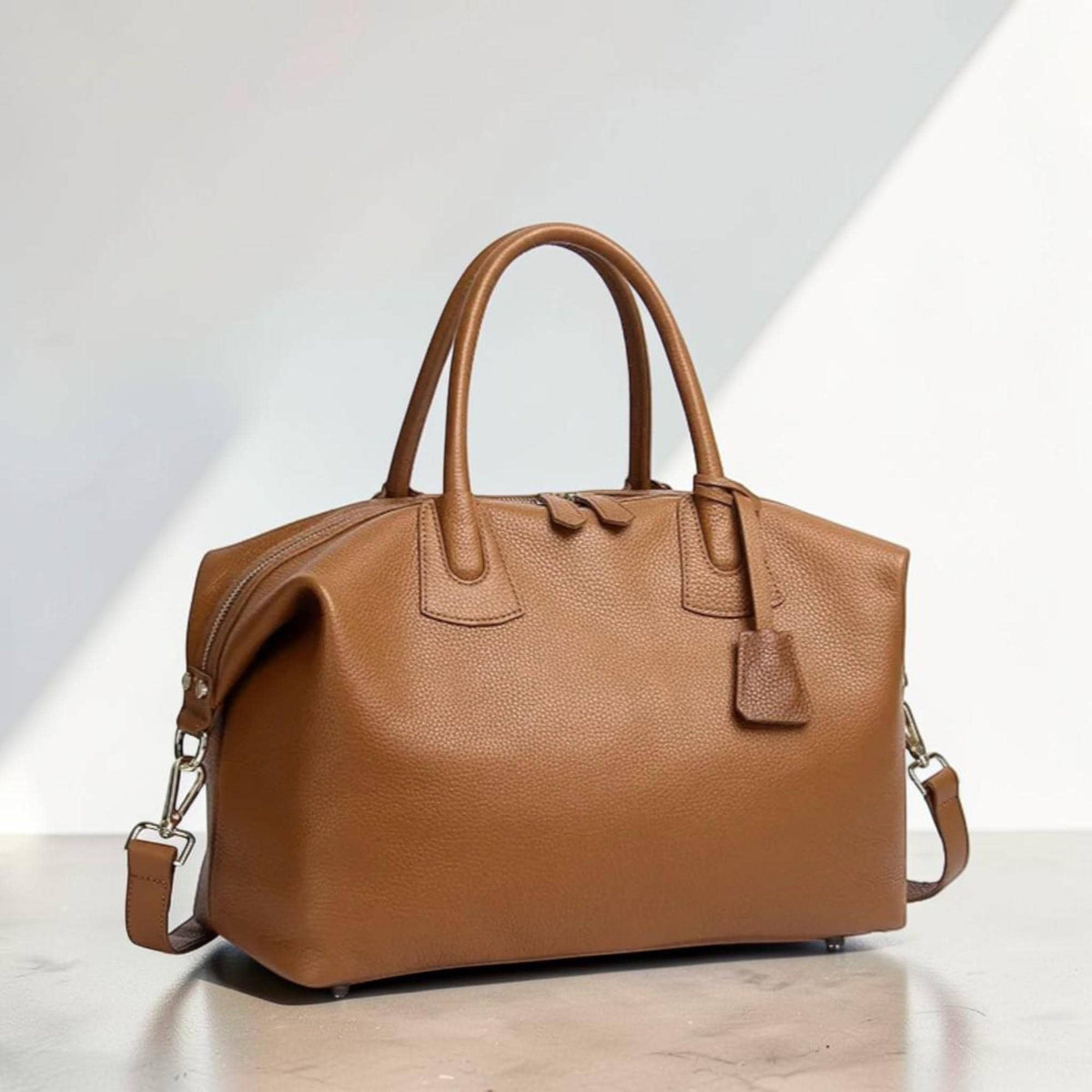 May include: A brown leather duffel bag with a top zipper closure, two top handles, and a detachable shoulder strap. The bag has a rectangular shape and a small leather tag attached to the zipper. The bag is made of textured leather.