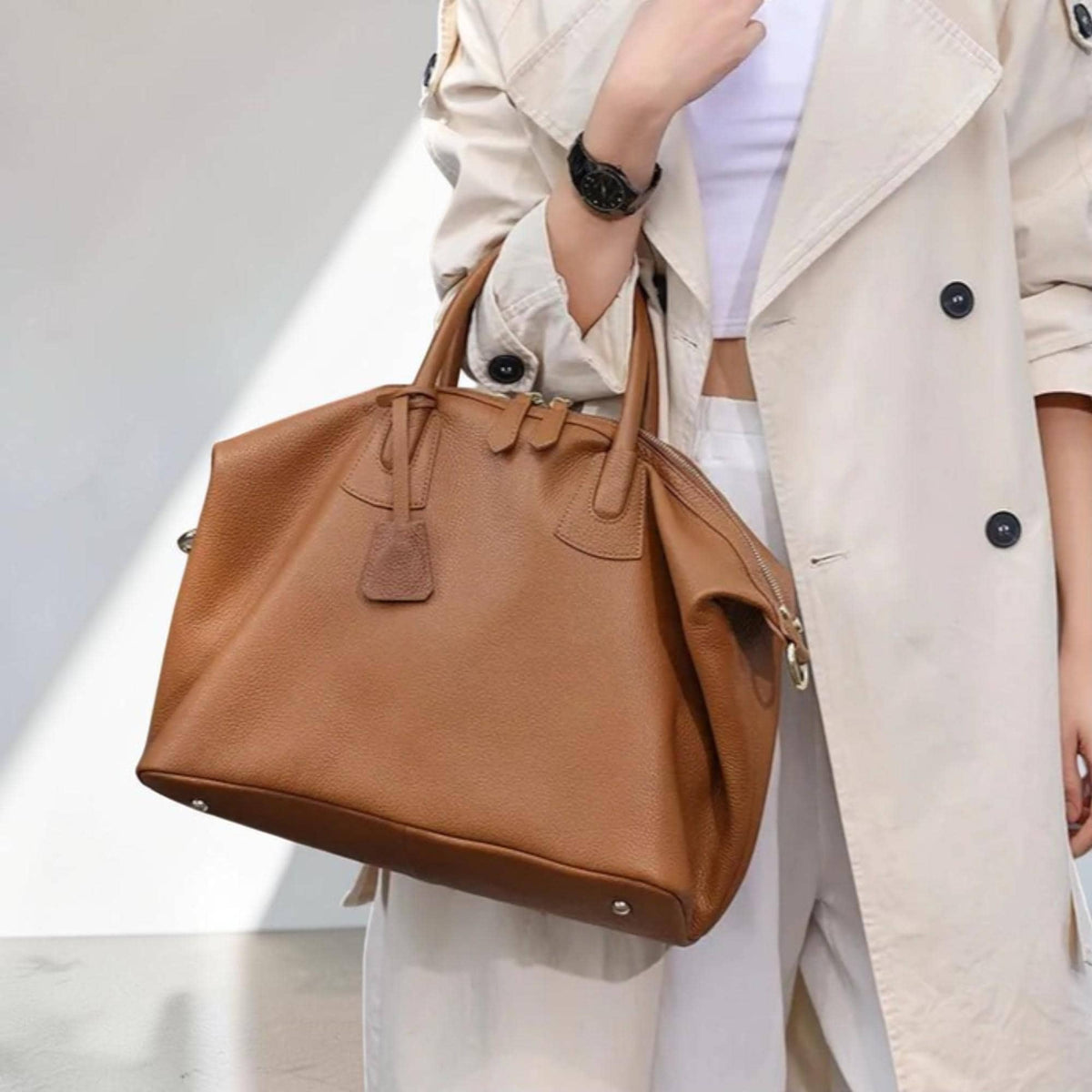May include: A brown leather handbag with a structured design. The bag features two top handles, a zipper closure, and a small leather tag. The bag is carried by a person wearing a light-colored trench coat.