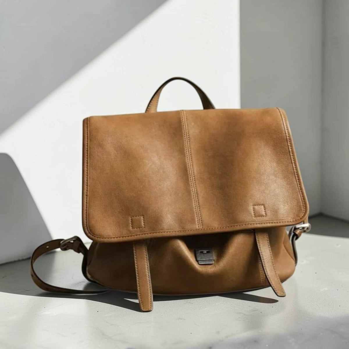 May include: A tan leather satchel with a flap closure, a top handle, and adjustable shoulder strap. The bag features a rectangular shape with a stitched vertical seam down the center and two buckle closures.