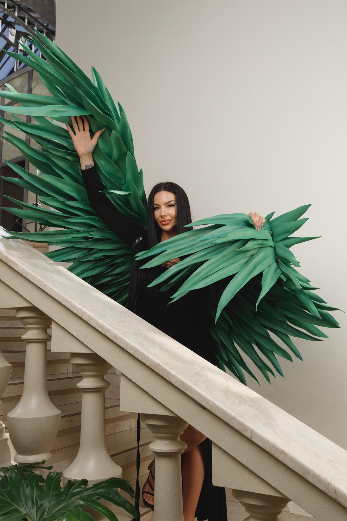 May include: Large, green, stylized leaf wings. The wings are held by a person wearing a black outfit, standing on a staircase with a white banister. The wings have a layered design and a textured appearance.