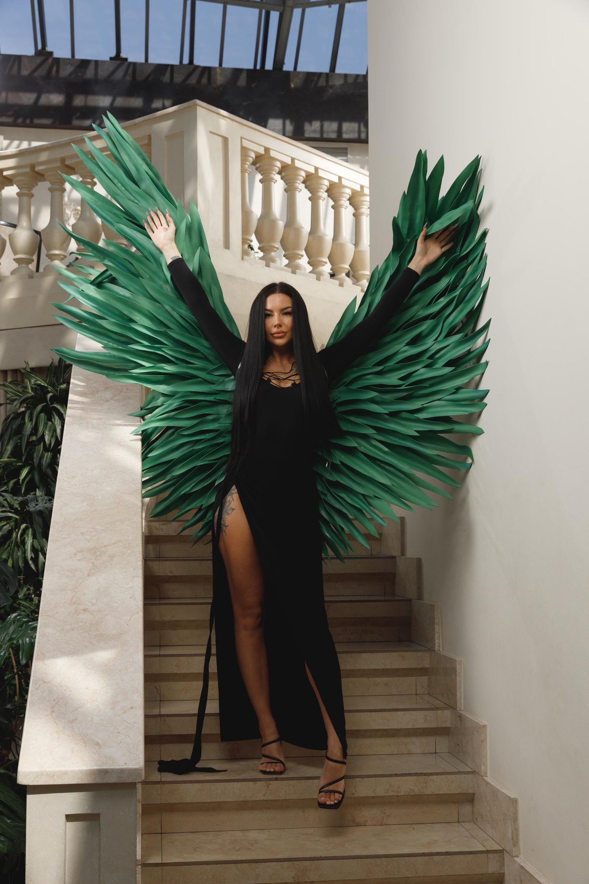 May include: A woman in a black dress with high slits and green angel wings stands on a staircase. The wings are large and made of layered, pointed green material. The dress has a low neckline and the woman is wearing black heels.